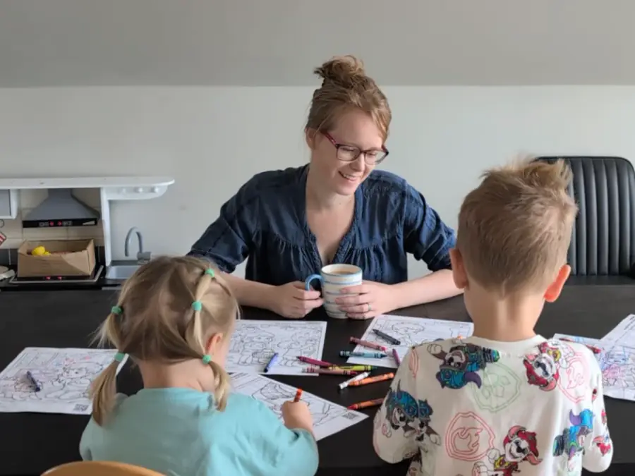 Sarah smiling over a mug at the kitchen table while her two children colour pages from the books