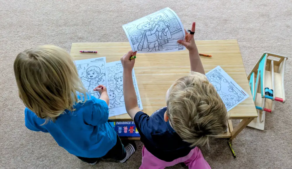 Two children seen from above at a low wooden table, one holding up a colouring page while the other colours beside her